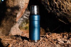 Thermos standing in a sunny forest. Vacuum travel flask close up. Camping concept, equipment for travel, adventure. photo