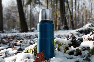 Hiking flask standing in winter forest, close-up. Vacuum blue thermos photo