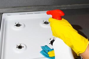 Female hand in rubber glove wiping the surface of gas stove, close-up. Woman washing kitchen apartment with cleaning spray photo