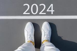 AI generated Number 2024 painted on asphalt, feet in sneakers standing in front of white horizontal line. Concept of the coming new year 2024. Top view, POV, close-up photo