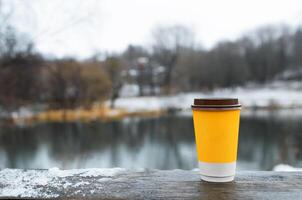 Cup of coffee standing on wooden table outdoors, copy space. Yellow paper glass with drink on winter background. photo