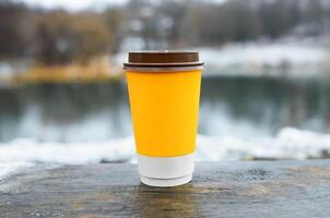 taza de café en pie en de madera mesa al aire libre, de cerca. amarillo papel vaso con bebida en invierno antecedentes. foto