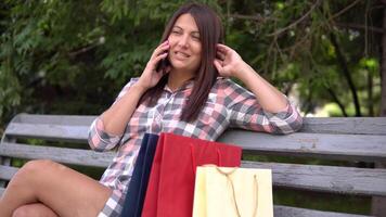 une magnifique fille après achats est assis dans le parc sur le banc et pourparlers sur le téléphone discuter le achats ayant une bien ambiance. 4k video