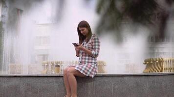 lindo menina dentro uma vestir senta perto uma fonte dentro a parque e usa uma Smartphone. lento movimento. hd video