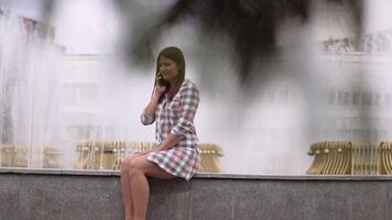 Beautiful girl in a dress sits near a fountain in the park and uses a smartphone. slow motion. video