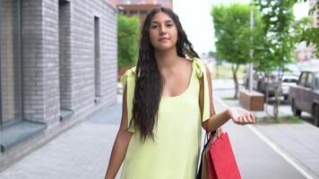 un hermosa niña con largo pelo en un amarillo vestir es caminando abajo el calle después compras. lento movimiento. hd video