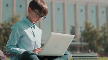 online opleiding, terug naar school, gelukkig schooljongen, leren lessen. schooljongen doet huiswerk gebruik makend van laptop terwijl zittend Aan de gras in de buurt de school. portret video