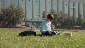Online education, Back to school, Happy schoolboy, Learn lessons. Schoolboy reads a book while sitting on the grass near the school. Move the camera to the right video