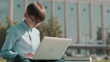 en línea educación, espalda a escuela, contento colegial, aprender lecciones colegial lo hace deberes utilizando ordenador portátil mientras sentado en el césped cerca el escuela. de cerca video