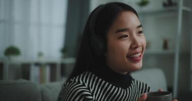 Selective focus,Happy young asian woman enjoy listening to the music with wireless headphones and sing while sitting drink coffee on sofa in living room at home, Leisure and lifestyle,Free time video