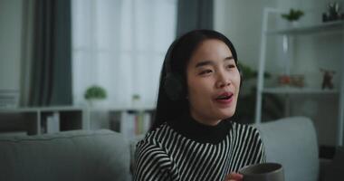 Selective focus,Happy young asian woman enjoy listening to the music with wireless headphones and sing while sitting drink coffee on sofa in living room at home, Leisure and lifestyle,Free time video