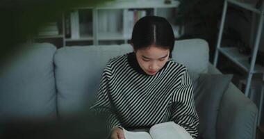 metraggio dolly sparato, felice giovane asiatico donna potabile mattina caffè o tè e lettura nel vivente camera a casa su fine settimana. tempo libero e stile di vita, libero tempo video