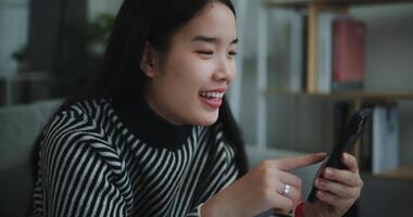 métrage côté vue de content Jeune asiatique femme asseoir sur canapé en portant café tasse et prendre plaisir prise selfies sur mobile téléphone à à la maison, gratuitement le temps, prends pause, sourire video