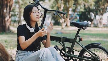 livsstil, utbildning, ungdom. Tonårs studerande i hörlurar med smartphone cykel Sammanträde i parkera, kvinna utseende på telefon skärm video