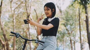 estilo de vida, educación, adolescencia. Adolescente estudiante en auriculares con teléfono inteligente bicicleta sentado en parque, hembra mira a teléfono pantalla video