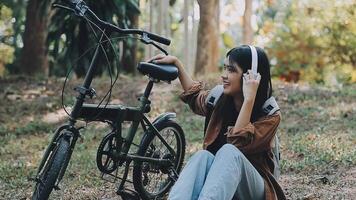 livsstil, utbildning, ungdom. Tonårs studerande i hörlurar med smartphone cykel Sammanträde i parkera, kvinna utseende på telefon skärm video