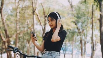 estilo de vida, educación, adolescencia. Adolescente estudiante en auriculares con teléfono inteligente bicicleta sentado en parque, hembra mira a teléfono pantalla video