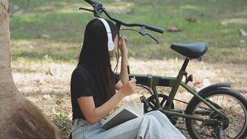 estilo de vida, educación, adolescencia. Adolescente estudiante en auriculares con teléfono inteligente bicicleta sentado en parque, hembra mira a teléfono pantalla video