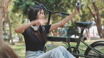 estilo de vida, educación, adolescencia. Adolescente estudiante en auriculares con teléfono inteligente bicicleta sentado en parque, hembra mira a teléfono pantalla video