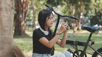 estilo de vida, Educação, adolescência. Adolescência aluna dentro fones de ouvido com Smartphone bicicleta sentado dentro parque, fêmea parece às telefone tela video