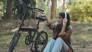 estilo de vida, educación, adolescencia. Adolescente estudiante en auriculares con teléfono inteligente bicicleta sentado en parque, hembra mira a teléfono pantalla video