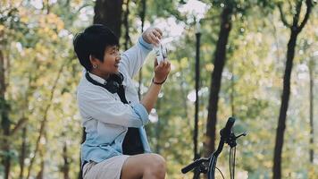 levensstijl, opleiding, adolescentie. tiener- leerling in hoofdtelefoons met smartphone fiets zittend in park, vrouw looks Bij telefoon scherm video