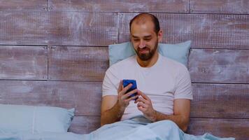 homme en utilisant téléphone et à la recherche à social médias contenu, souriant et en riant dans chambre à coucher. Zoom en dehors coup video