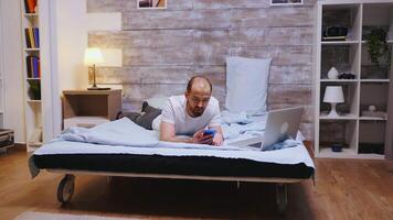 Man in pajamas browsing on smartphone while working on laptop before bedtime video