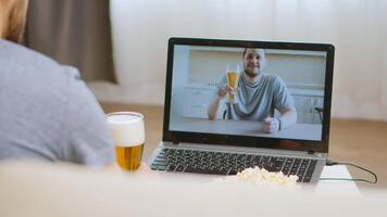 Back view of happy man on video call with his friend drinking beer during coronavirus isolation.