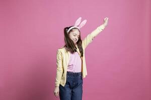 Positive enthusiastic girl fooling around and dancing on camera, wearing bunny ears in studio and showing dance moves. Young toddler acting cheerful and excited about easter celebration. photo