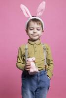 Young small child smiling in studio and holding a cute pink rabbit toy, celebrating easter holiday with handmade painted decoration. Joyful little boy wearing bunny ears and poses on camera. photo