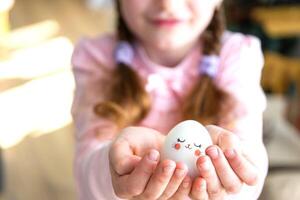 A cute girl with pink bunny ears makes an Easter craft - decorates an egg in the form of a unicorn with rhinestones, horn, flowers in the interior of a house with plants. photo