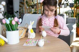 A cute girl with pink bunny ears makes an Easter craft - decorates an egg in the form of a unicorn with rhinestones, horn, flowers in the interior of a house with plants. photo