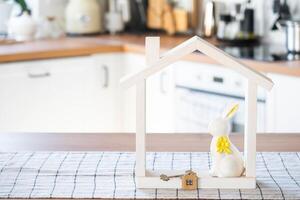 Key and tiny house of cozy home with Easter decor with rabbit and eggs on table of kitchen. Building, design, project, moving to new house, mortgage, insurance, rent and purchase real estate photo