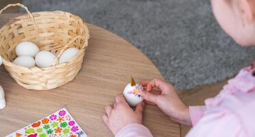 A cute girl with pink bunny ears makes an Easter craft - decorates an egg in the form of a unicorn with rhinestones, horn, flowers in the interior of a house with plants. photo