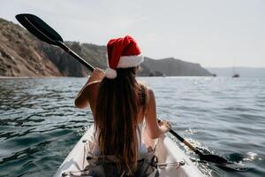 Woman in kayak back view. Happy young woman in Santa hat floating in kayak on calm sea. Summer holiday vacation and cheerful female people relaxing having fun on the boat. photo