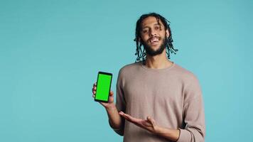 Happy man pointing towards green screen mobile phone, showing thumbs up sign. Cheerful person presenting chroma key cellphone, doing positive gesturing, isolated over studio background, camera A video