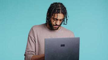 mitten östra man inmatning data på bärbar dator medan stående i blå studio bakgrund. bipoc anställd skriver på anteckningsbok tangentbord, fokuserade på lösning projekt uppgifter, kamera b video