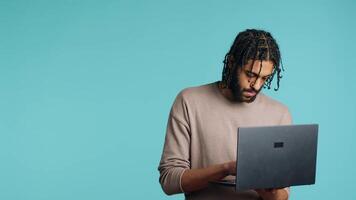 mitten östra man inmatning data på bärbar dator medan stående i blå studio bakgrund. bipoc anställd skriver på anteckningsbok tangentbord, fokuserade på lösning projekt uppgifter, kamera en video