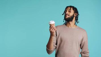 Man enjoys fresh coffee from disposable paper cup early in the morning to wake up and be energized. Person drinks hot beverage from brown recycled takeaway cappuccino cup, studio background, camera A video