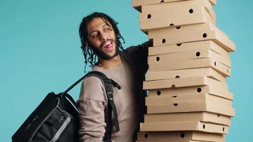 Portrait of smiling pizza delivery man holding takeaway orders for customer. Upbeat BIPOC courier with lunch boxes in arms and thermal food backpack, isolated over blue studio background, camera A video