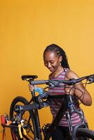 Active healthy female securing bike on repair-stand for repairing against yellow background. Image showing sporty black woman adjusting bicycle height, ready for yearly maintenance. photo