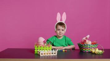 dolce poco bambino mettendo soffice coniglietto orecchie su il suo testa e sorridente su telecamera, contento innocente ragazzo seduta a un' tavolo con arte e mestiere forniture per decorare Pasqua uova. primavera vacanza. telecamera b. video