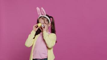 adorable peu fille en jouant jeter un œil une huer Jeu dans studio, montrant sa fabriqués à la main coloré Pâques des œufs contre rose Contexte. de bonne humeur espiègle enfant avec lapin oreilles tromper autour. caméra un. video