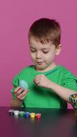 Vertical Video Joyful small kid painting eggs for easter holiday festivity in studio, using watercolor and art supplies. Smiling preschooler coloring festive ornaments in preparation for sunday. Camera A.