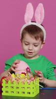 Vertical Video Joyful smiling boy decorating baskets and arrangements for easter sunday celebration, putting plastic grass and fake flowers to craft festive decorations. Happy toddler with bunny ears. Camera A.