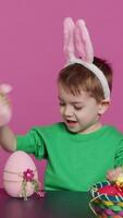 verticale vidéo joyeux Jeune enfant en jouant autour avec de fête peint décorations, montrant une lapin jouet et une rose Oeuf dans de face de caméra. souriant petit garçon avec lapin oreilles ayant amusement avec ornements. caméra un. video