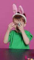 Vertical Video Smiling cute kid playing peek a boo game with painted colorful eggs, having fun with easter decorations and arrangements on camera. Enthusiastic joyful boy being excited about holiday. Camera A.