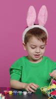 Vertical Video Smiling jolly preschooler painting eggs and ornaments for easter festivity preparations, using art and craft materials to decorate festive arrangements. Young boy having fun with tie dye. Camera A.