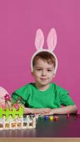 Sweet little child putting fluffy bunny ears on his head and smiling on camera, happy innocent boy sitting at a table with art and craft supplies to decorate easter eggs. Spring holiday. Camera B. video